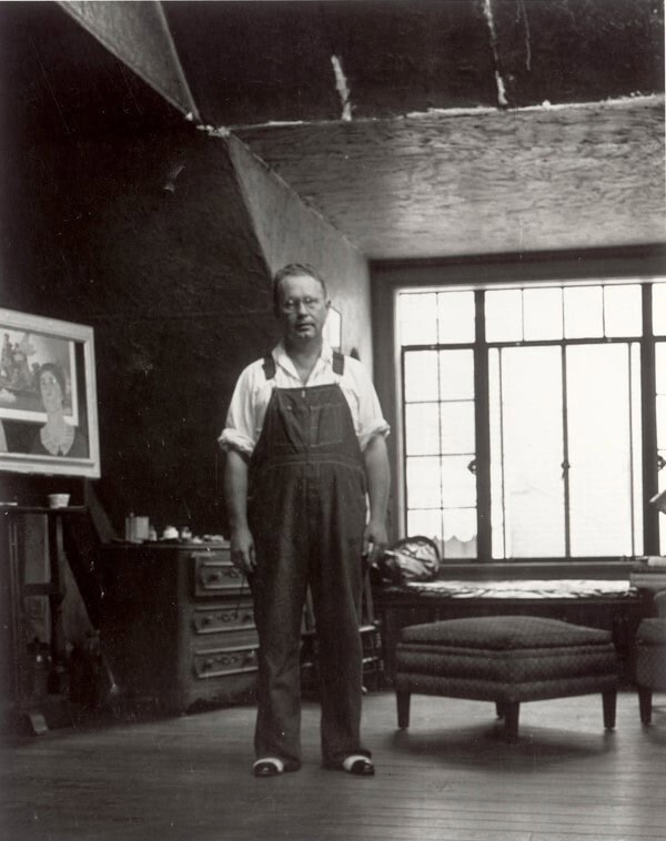 A middle aged man wearing overalls and glasses stands in a tidy, minimally furnished room.